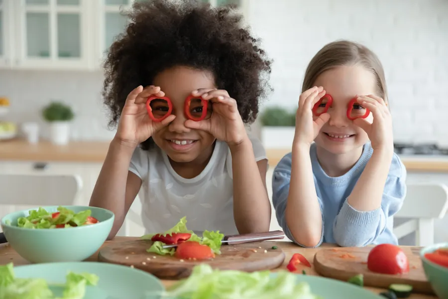 two girls in kitchen making salad hold up veggie slices on face